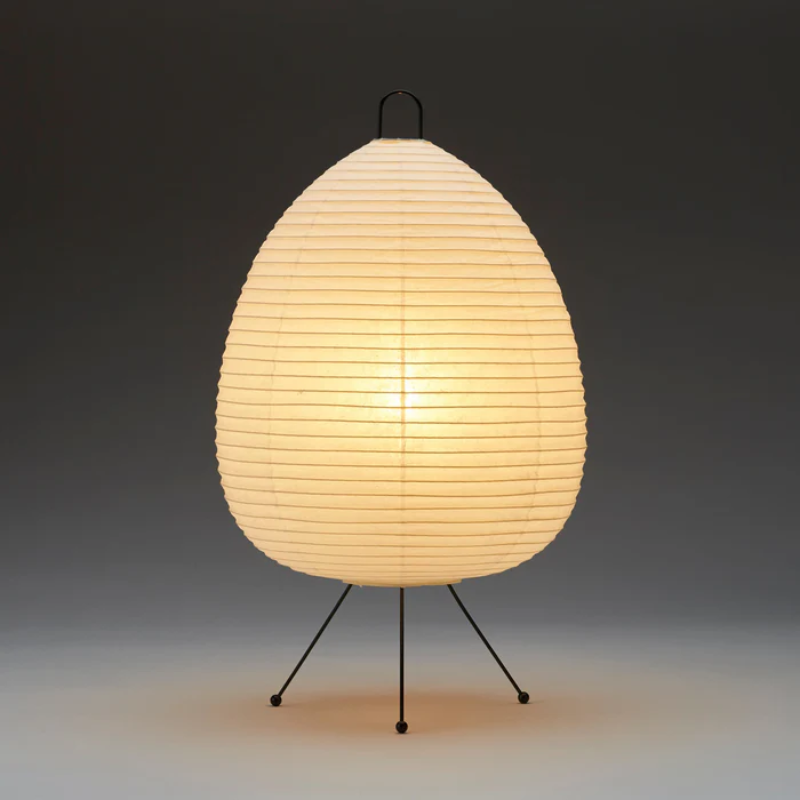 The Asano Donguri Lamp A120 has a white rice paper shade and black metal legs. Lamp turned on with warm glow.