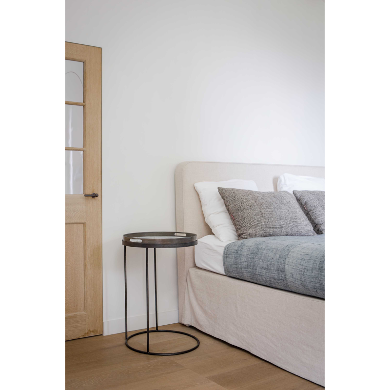 The Round Tray Side Table from Ethnicraft beside a bed in a primary bedroom.