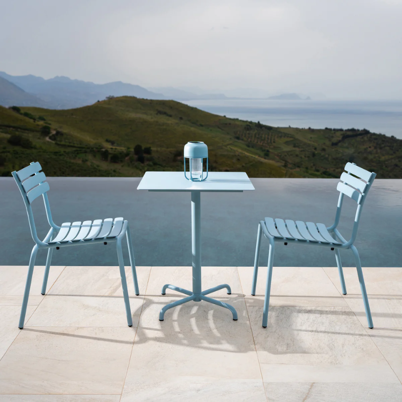 The ice blue Pico Outdoor Square Cafe Table 4-Star Base by Houe with chairs set with a scenic background of mountains and sea.