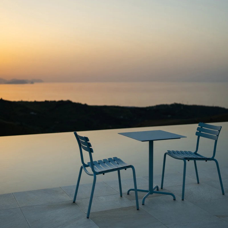 The ice blue Pico Outdoor Square Cafe Table 4-Star Base by Houe Two blue chairs and a table on a patio overlooking a sunset over water.