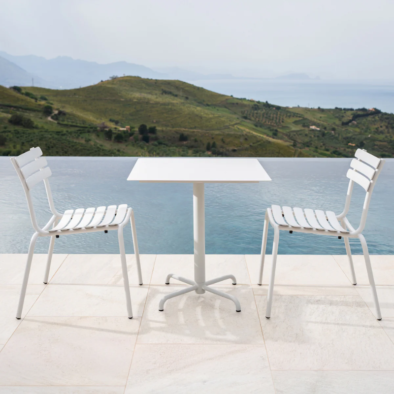 The muted white Pico Outdoor Square Cafe Table 4-Star Base by Houe with chairs has a scenic view of hills and water.