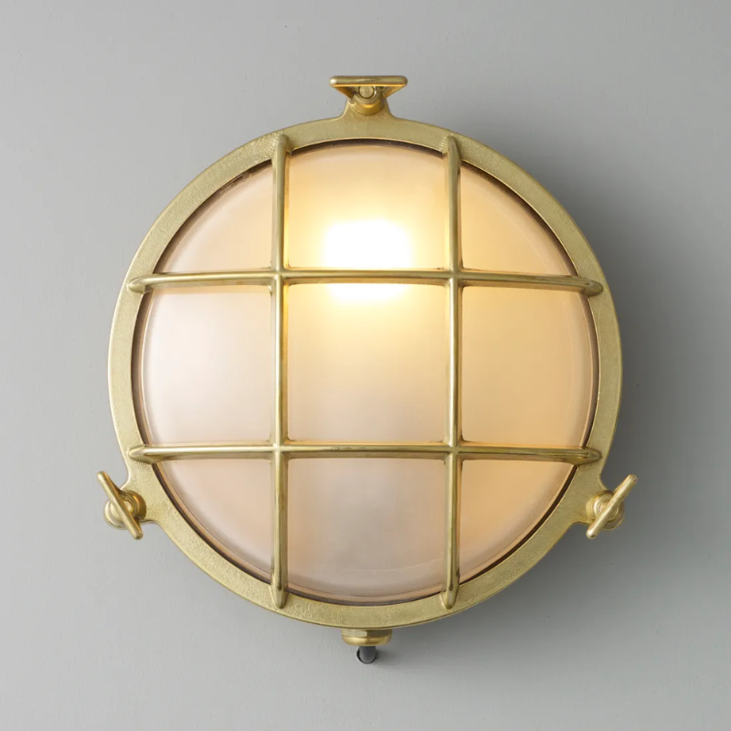 The medium Brass Bulkhead Outdoor Wall Sconce from Original BTC in polished brass in a studio turned on.
