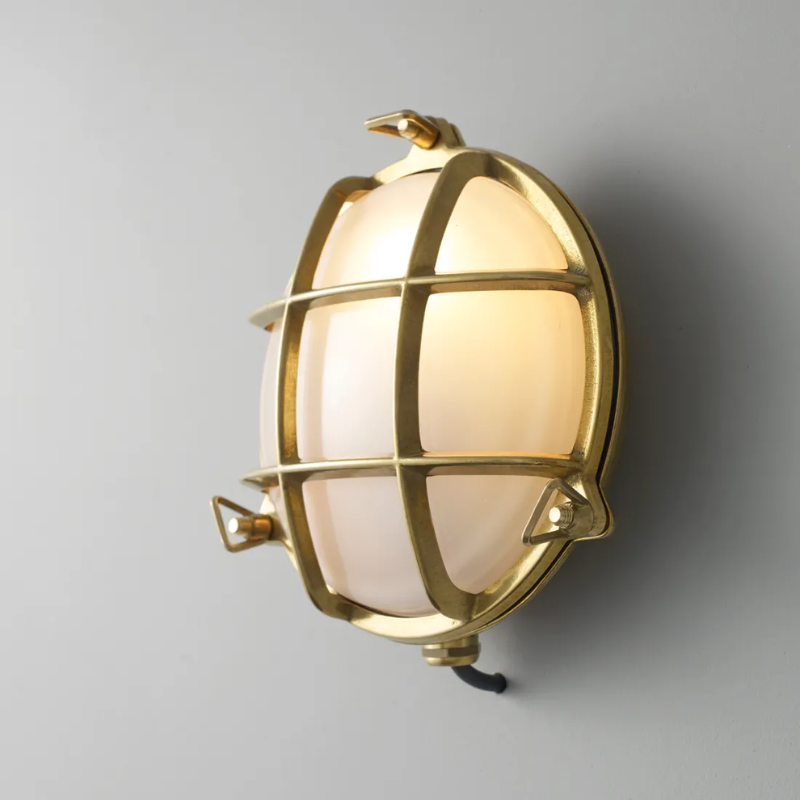 The small Brass Bulkhead Outdoor Wall Sconce from Original BTC in polished brass in a studio from an angle turned on