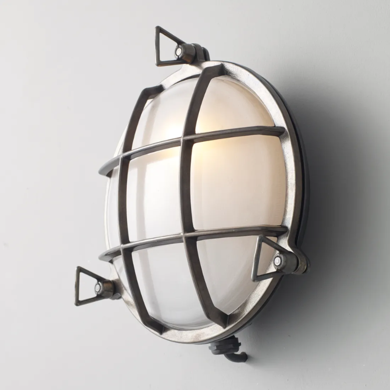 The medium Brass Bulkhead Outdoor Wall Sconce from Original BTC in weathered brass in a studio from an angle turned on.