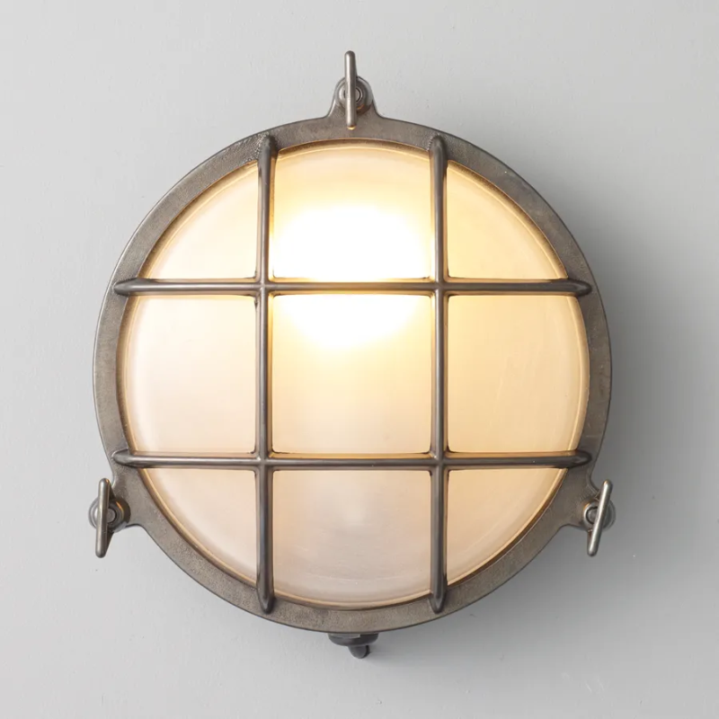 The medium Brass Bulkhead Outdoor Wall Sconce from Original BTC in weathered brass in a studio turned on.