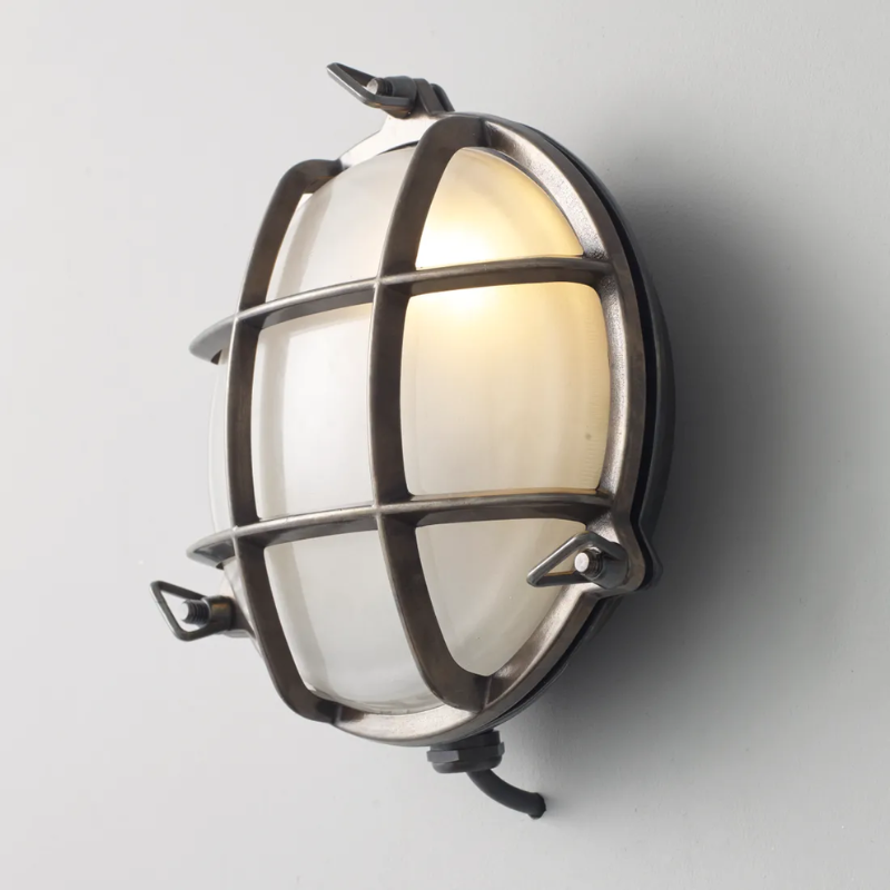 The small Brass Bulkhead Outdoor Wall Sconce from Original BTC in weathered brass in a studio from an angle turned on.