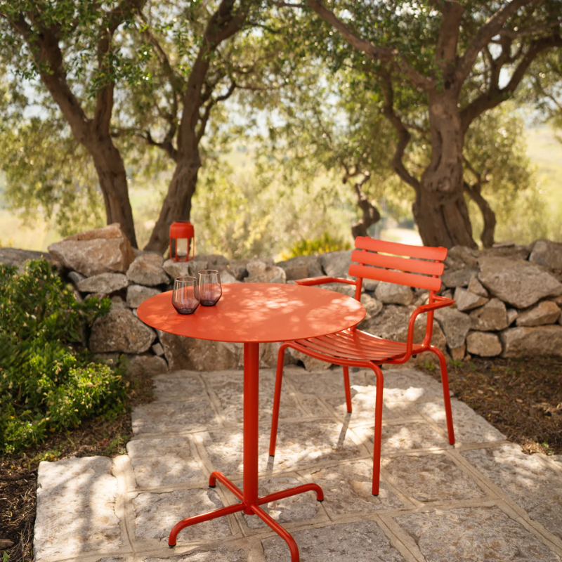 The Pico Round Cafe Table 4-Star Base by Houe is in an outdoor area with a chair on a stone patio with trees and rocks. all-groups