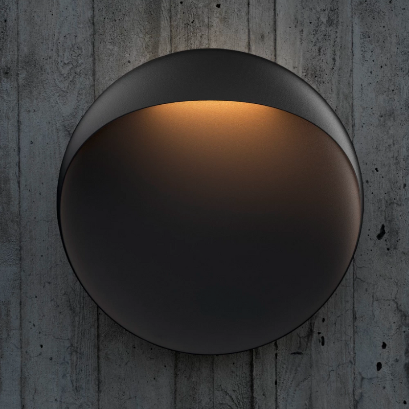 The Flindt Wall is distinguished by a light egress from an opening in the top that emits light down the front of the fixture, creating a subtle, glowing ring of reflected light on the transition between the concave and convex shapes. The oval shape is lifted off the mounting surface, giving the fixture a floating appearance.