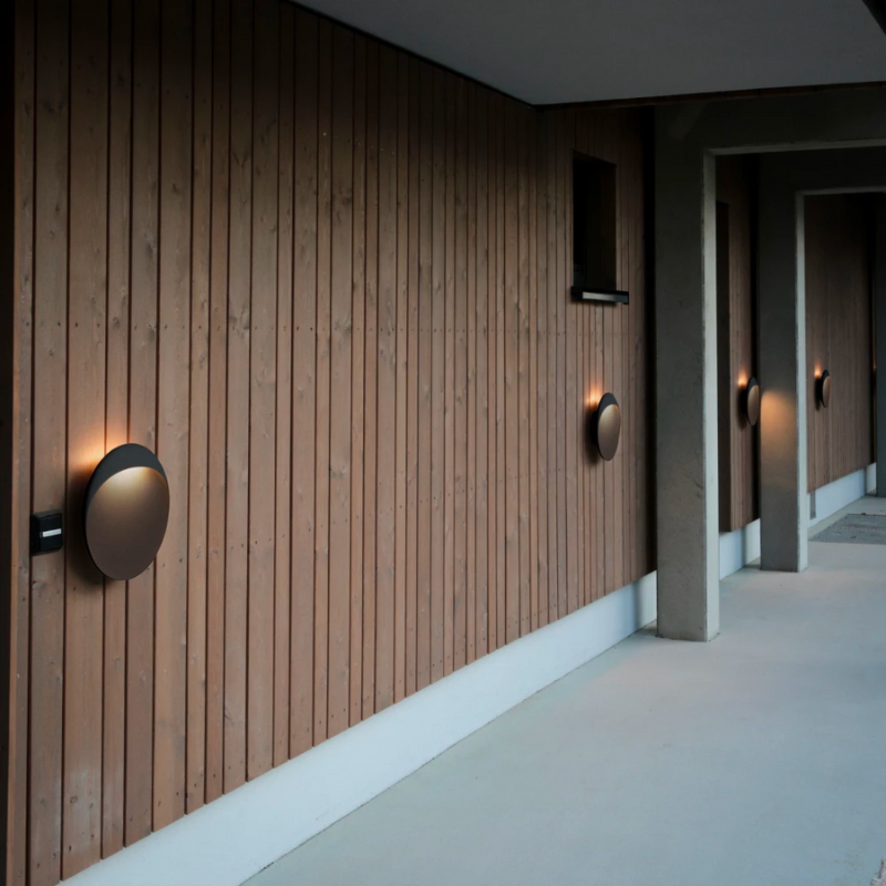 The Flindt Wall is distinguished by a light egress from an opening in the top that emits light down the front of the fixture, creating a subtle, glowing ring of reflected light on the transition between the concave and convex shapes. The oval shape is lifted off the mounting surface, giving the fixture a floating appearance.