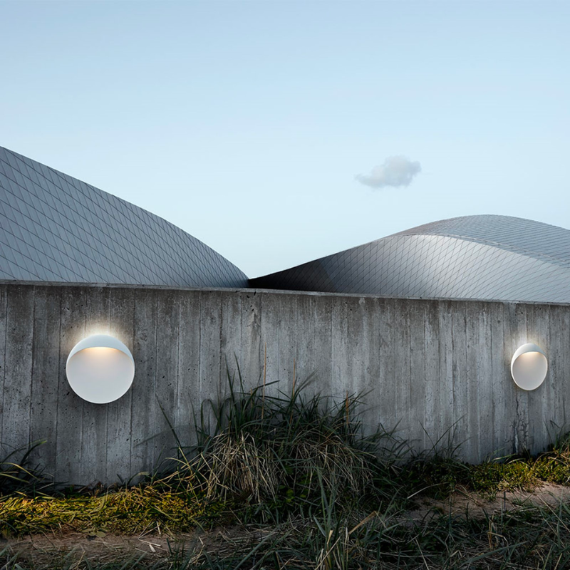 The Flindt Wall is distinguished by a light egress from an opening in the top that emits light down the front of the fixture, creating a subtle, glowing ring of reflected light on the transition between the concave and convex shapes. The oval shape is lifted off the mounting surface, giving the fixture a floating appearance.