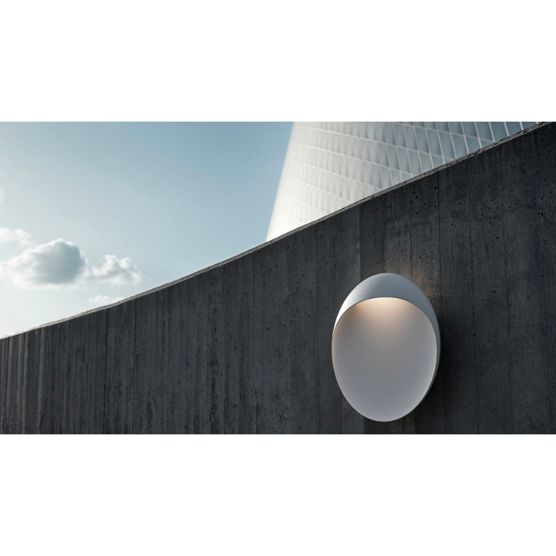 The Flindt Wall is distinguished by a light egress from an opening in the top that emits light down the front of the fixture, creating a subtle, glowing ring of reflected light on the transition between the concave and convex shapes. The oval shape is lifted off the mounting surface, giving the fixture a floating appearance.