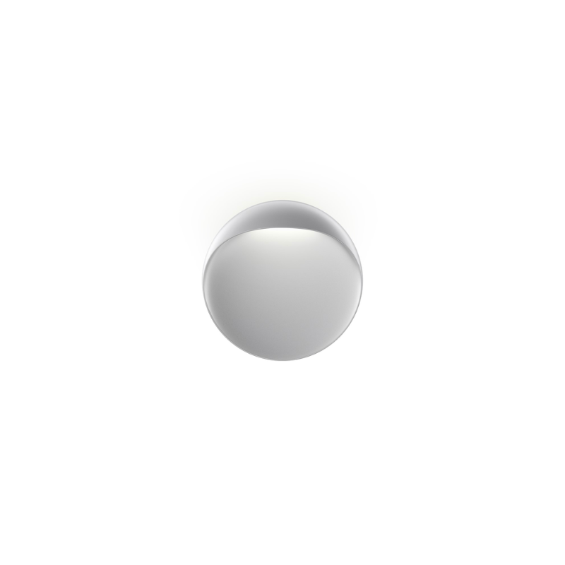 The Flindt Wall is distinguished by a light egress from an opening in the top that emits light down the front of the fixture, creating a subtle, glowing ring of reflected light on the transition between the concave and convex shapes. The oval shape is lifted off the mounting surface, giving the fixture a floating appearance.