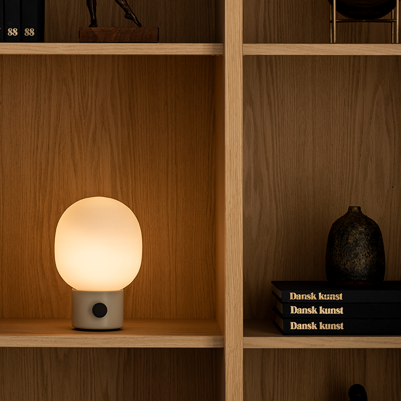 The JWDA Portable Table Lamp from Audo Copenhagen in alabaster white in a bedroom on a shelf without a power source.