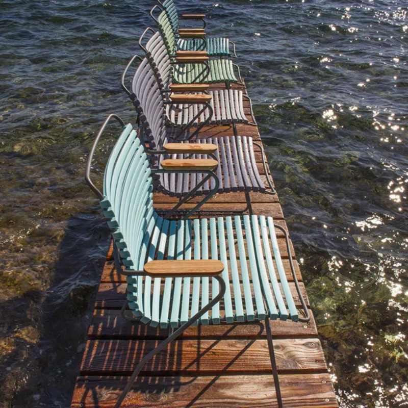 The Click Outdoor Dining Chair by Houe is shown in multiple color options on a wooden dock overlooking the sea.