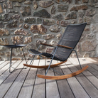 The Click Outdoor Rocking Chair by Houe on a wooden deck beside a stone wall background.