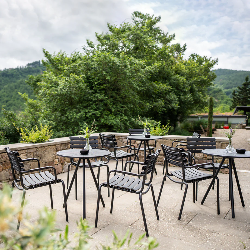 The black ReClips Outdoor Dining Chair Aluminium by Houe in an outdoor seating area with tables on a stone patio with greenery.