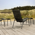The ReClips Outdoor Footrest by Houe with a lounge chair on a wooden deck with grass and trees.