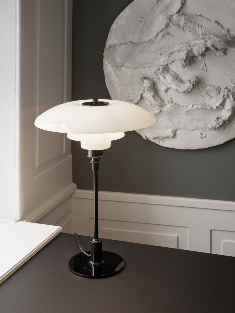 The PH 3½-2½ glass table lamp is designed based on the principle of a reflective three-shade system, which directs the majority of the light downwards. The shades are made of hand blown opal three-layer glass, which is glossy on top and sandblasted matte on the underside, giving a soft and diffused light distribution. Designed by Poul Henningsen.